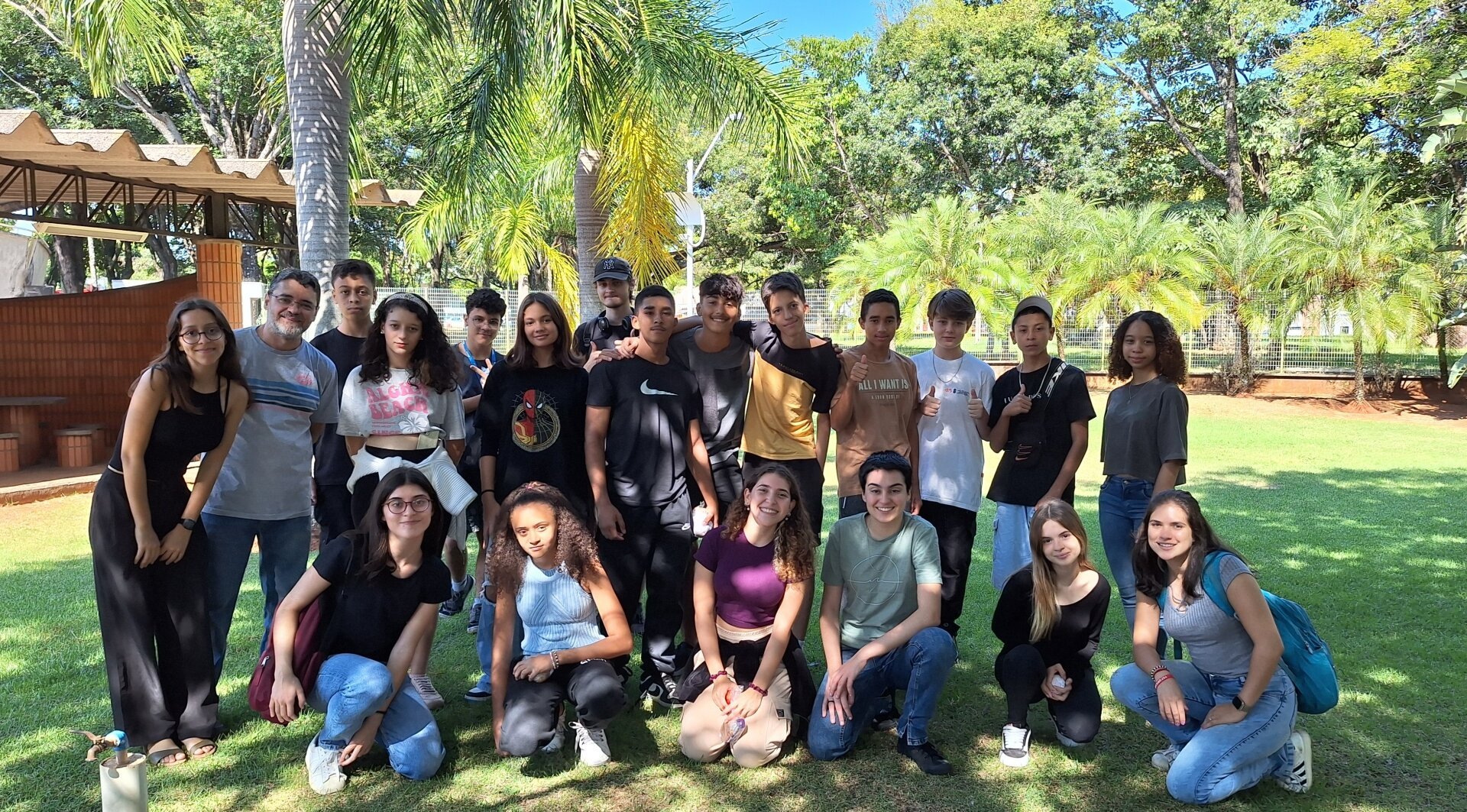 Foto de adolescentes e jovens, em gramado e num dia ensolarado. Alguns estão ajoelhados em fileira a frente e os demais perfilados, em pé. Entre eles, há um homem mais velho. Todos estão sorrindo.